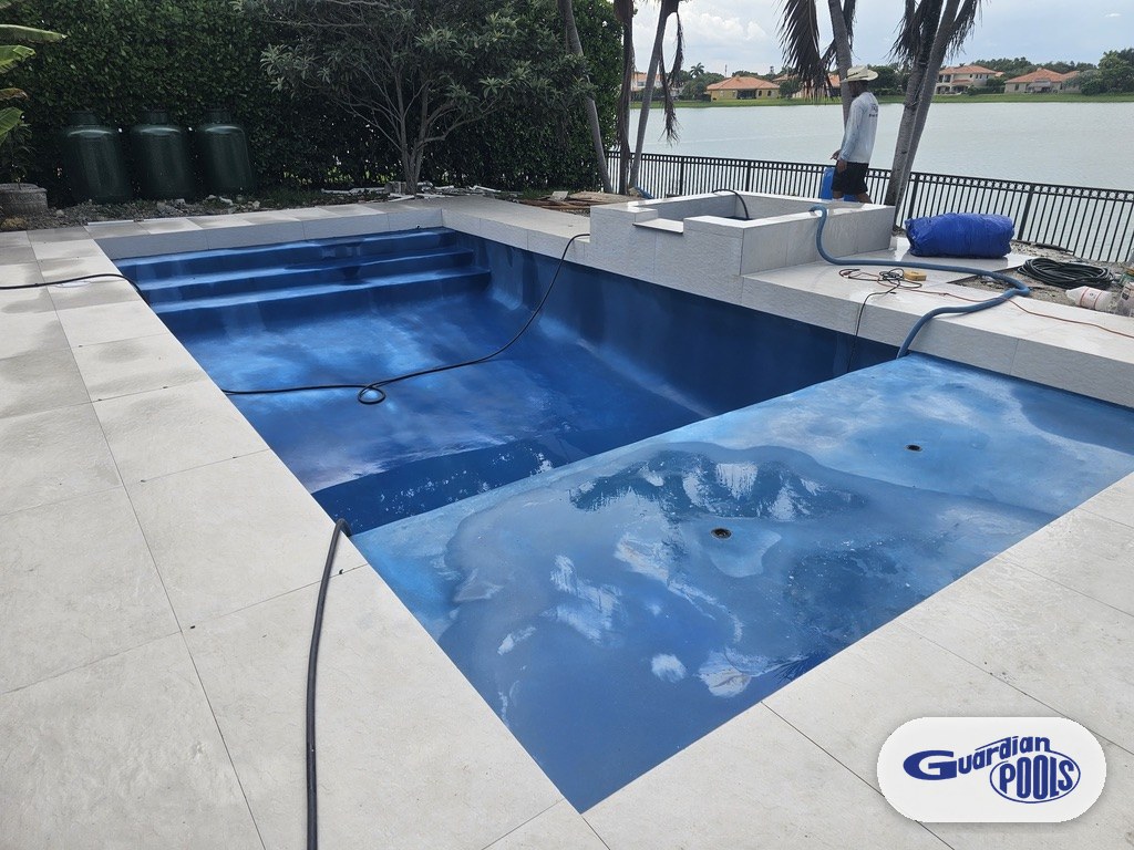 Pool and raised spa mid-construction with new dark blue finish and white paver deck, filling with water